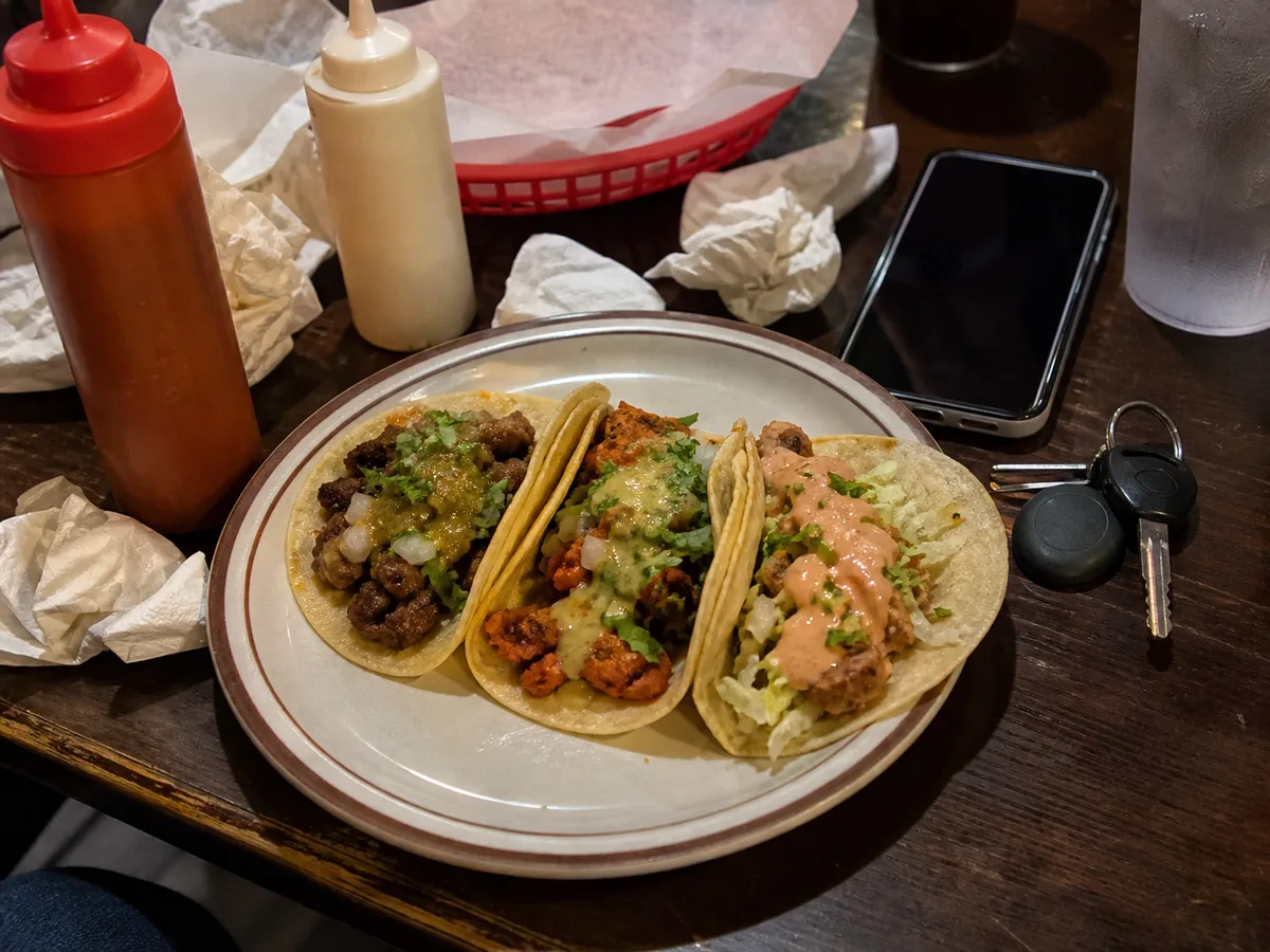 Tacos surrounded by napkins, a sauce bottle, a phone, and keys on a dim table