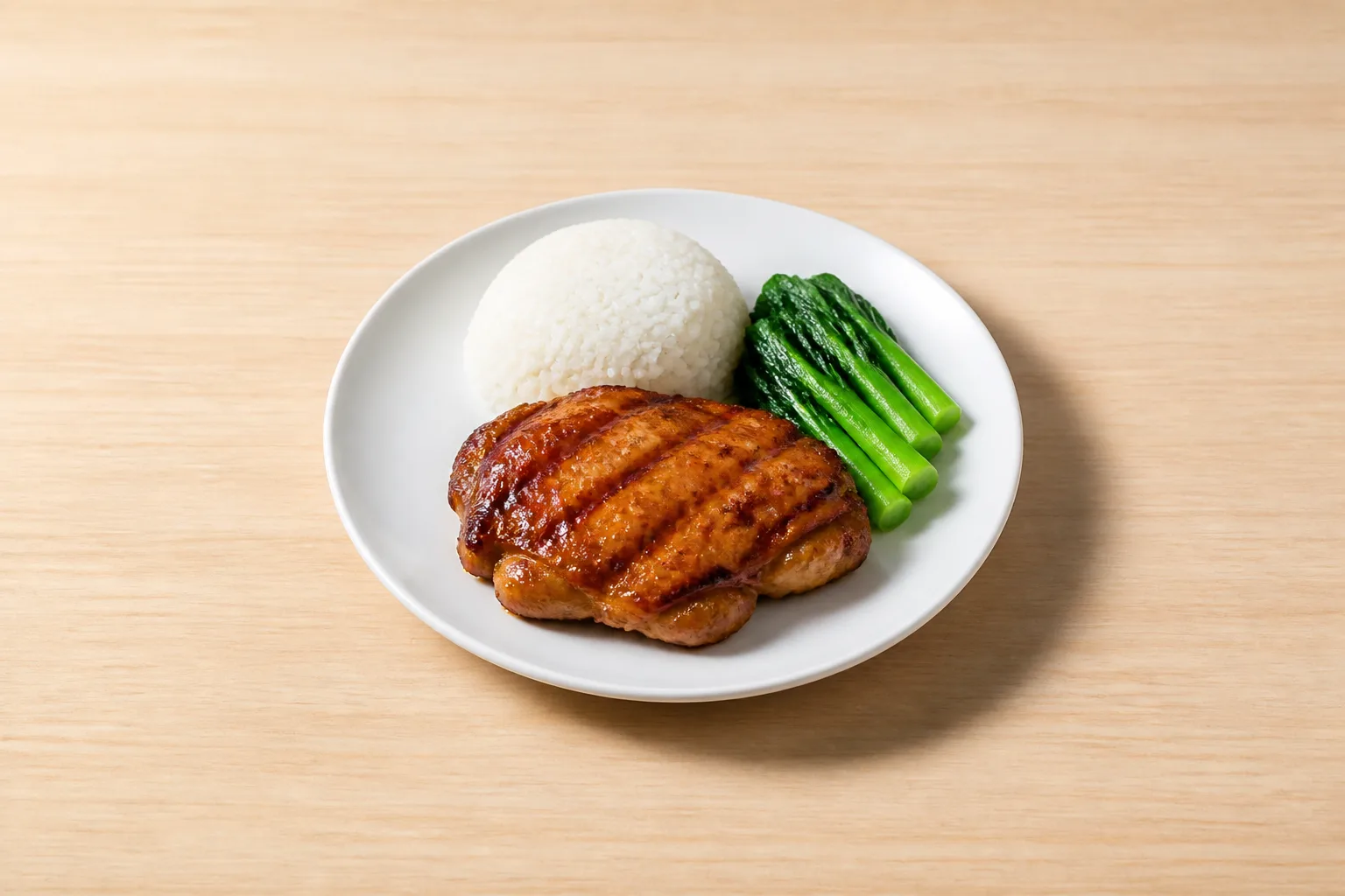 A plated grilled chicken thigh with rice and greens sits centered on a pale wooden table, lit by soft side light that leaves a pale, soft-edged shadow beside the plate