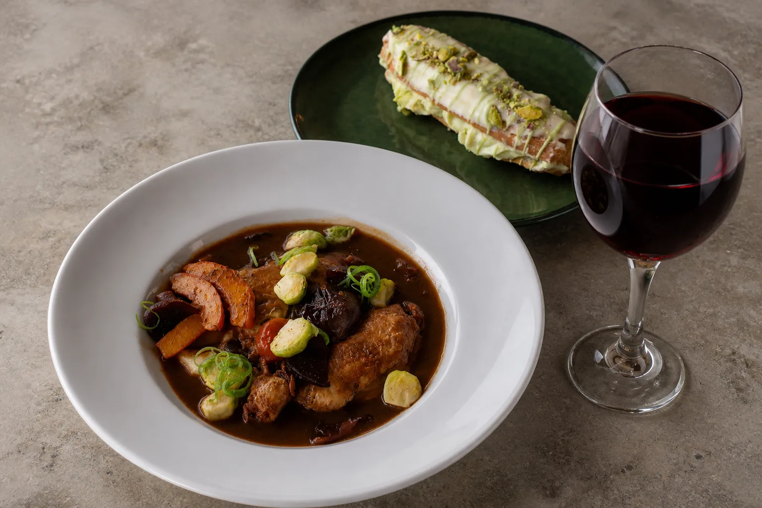 A plated coq au vin in a wide white bowl, the chicken and glazed vegetables sitting in a dark red wine reduction, beside a pistachio eclair on a dark green plate and a stemmed glass of red wine, all on a worn pale grey stone surface under one large soft side light