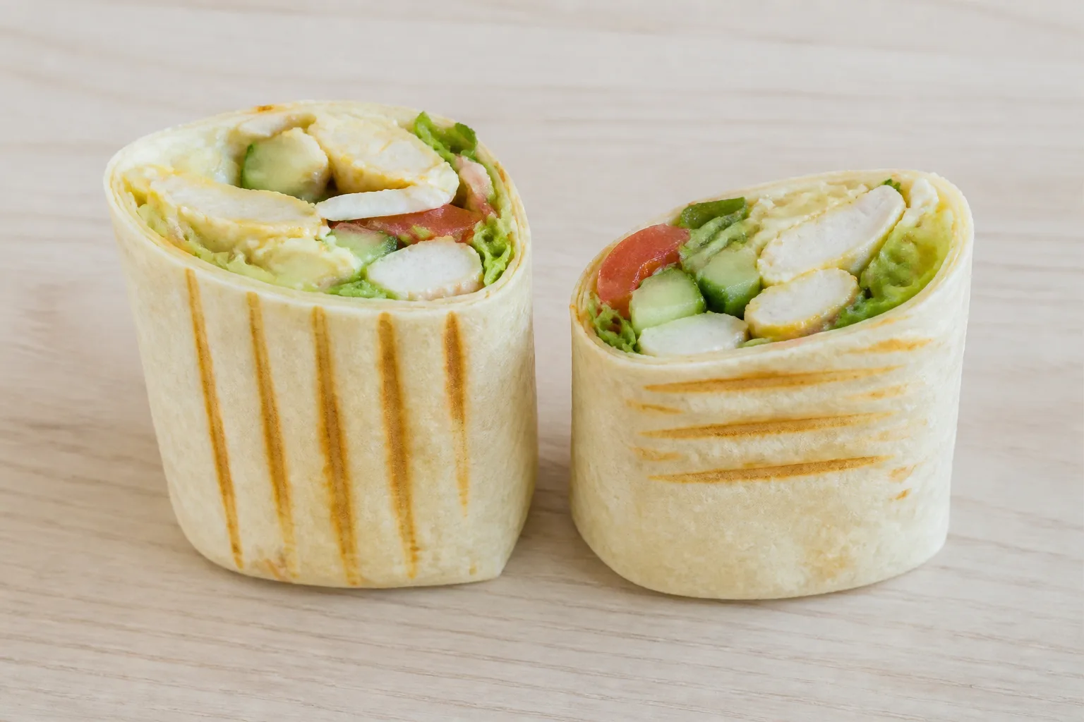 A burrito cut in half with both halves standing upright on their folded ends, cut faces to the camera showing avocado, tomato and cucumber in cross section, on a plain light wood surface against an empty pale background