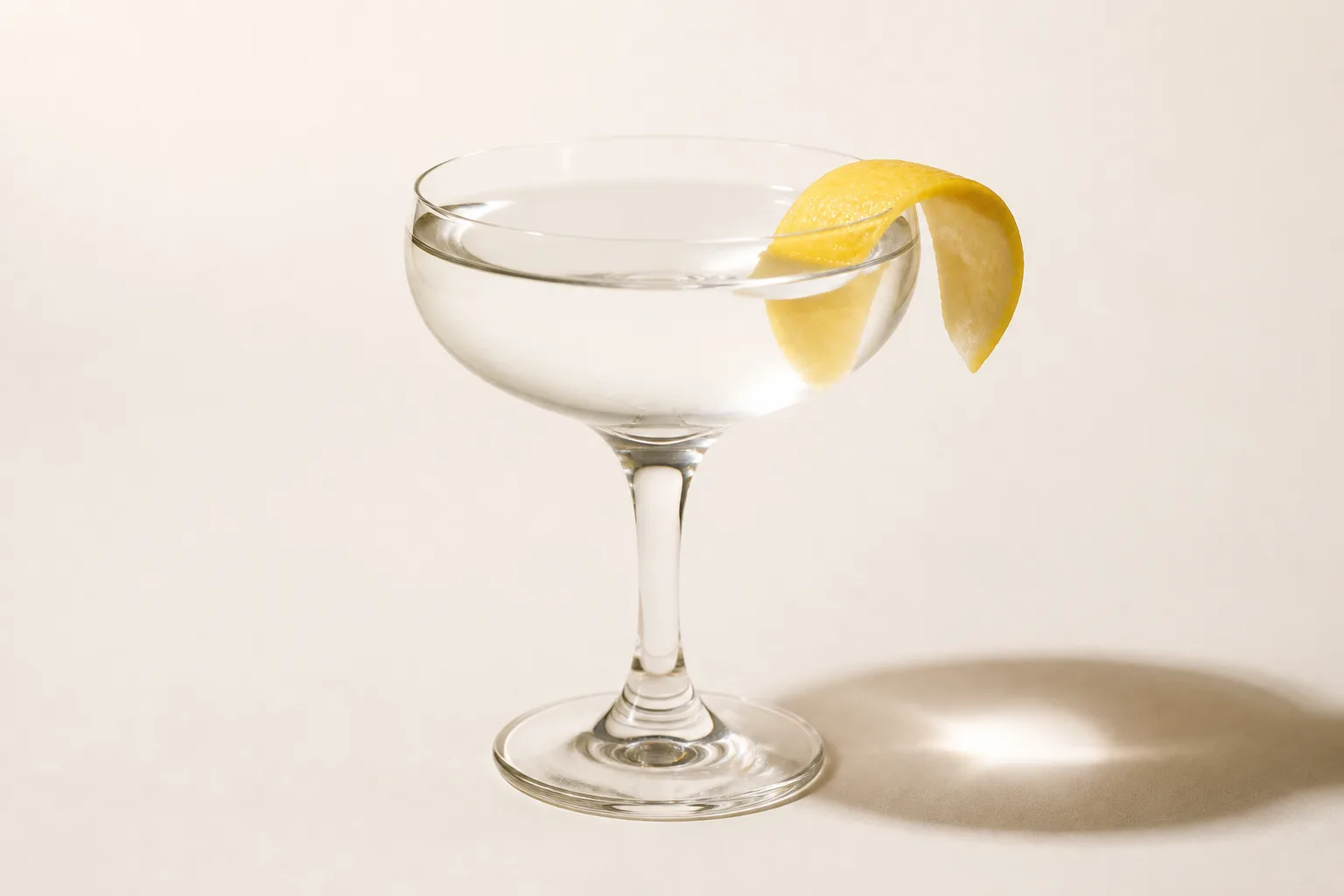 A clear cocktail in a coupe glass with a citrus twist garnish resting on the rim, shot straight on against an empty pale background