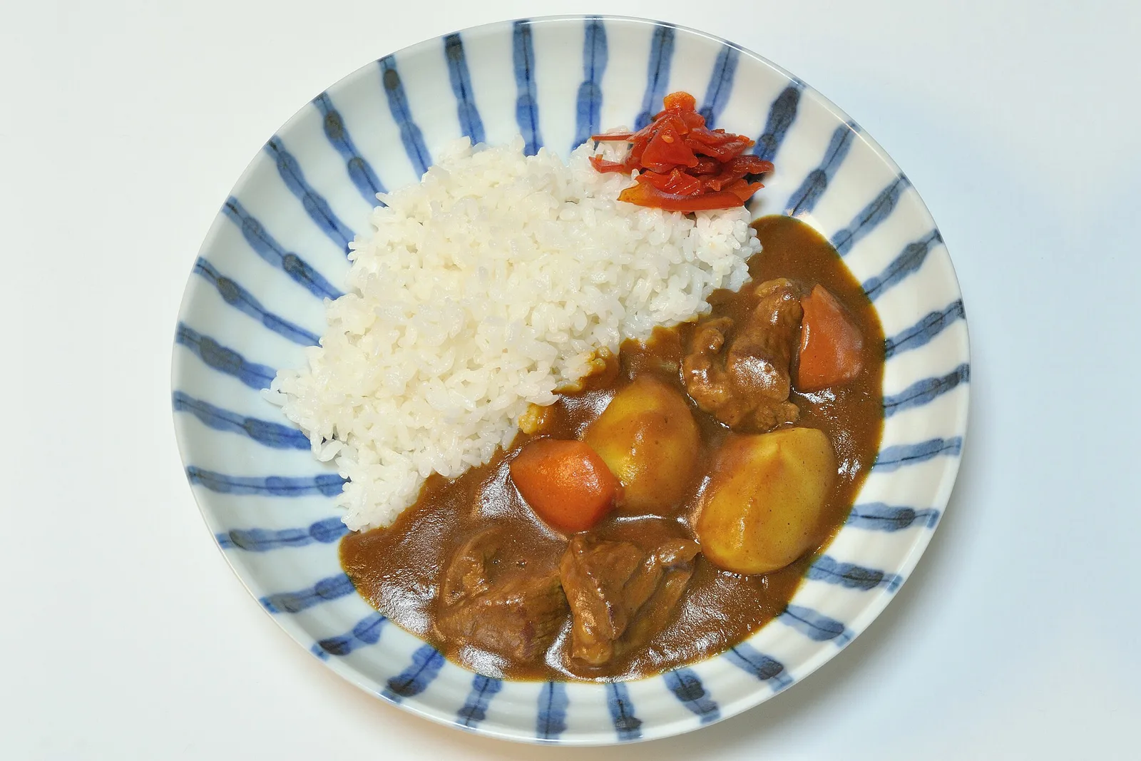 A plate of Japanese beef curry: mounded white rice on one side, a thick sauce with beef, potato and carrot on the other, and a heap of red pickle, on a blue-striped plate