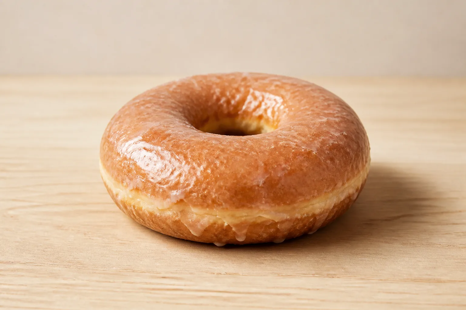 A single glazed ring donut with a glossy reflective glaze, shot from a three-quarter angle on a plain light wood surface against an empty pale background