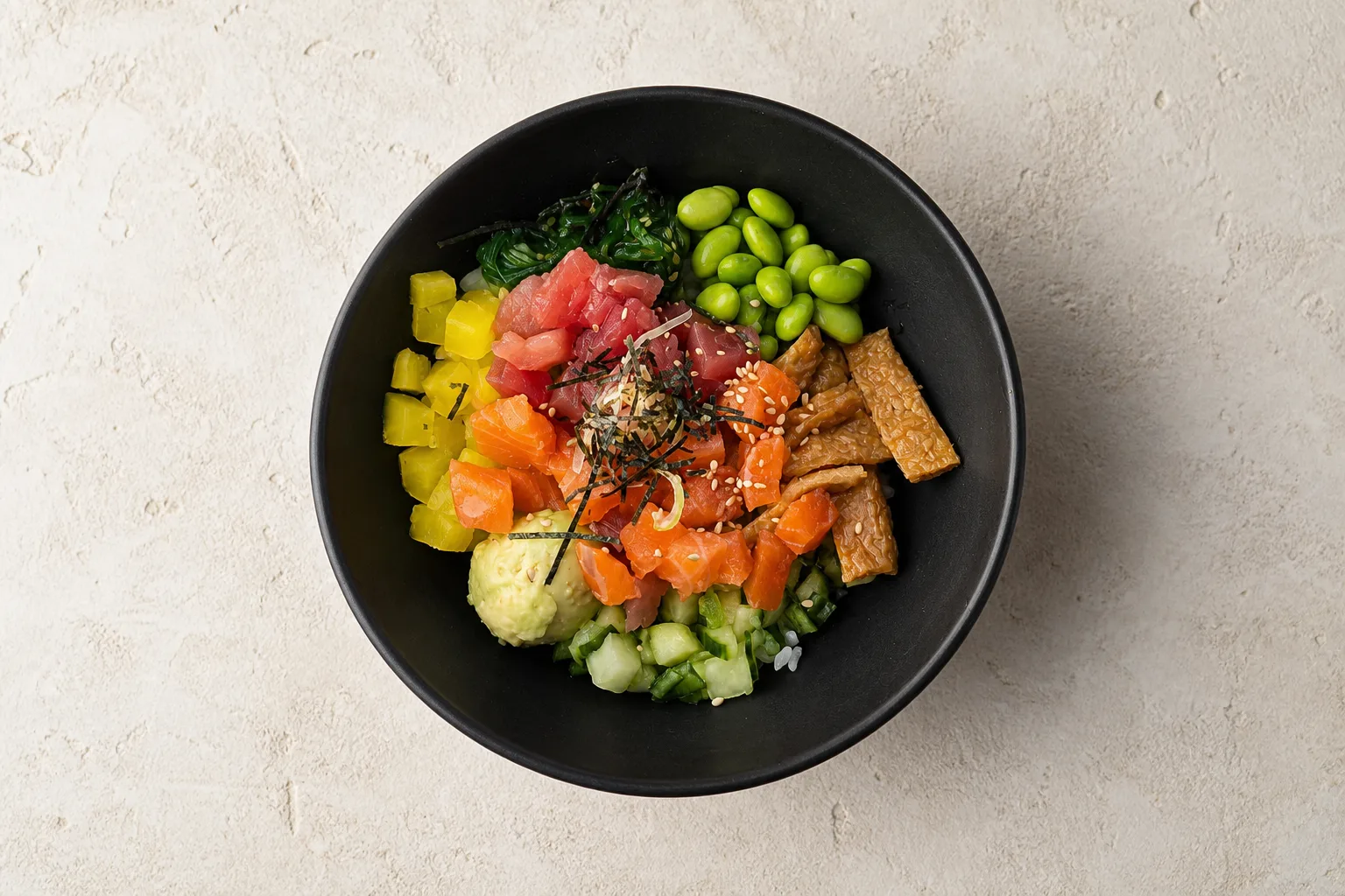 A poke bowl with cubed salmon and tuna, edamame, pickled radish, cucumber, fried tofu and nori arranged in sections over rice, shot from directly overhead against an empty pale background