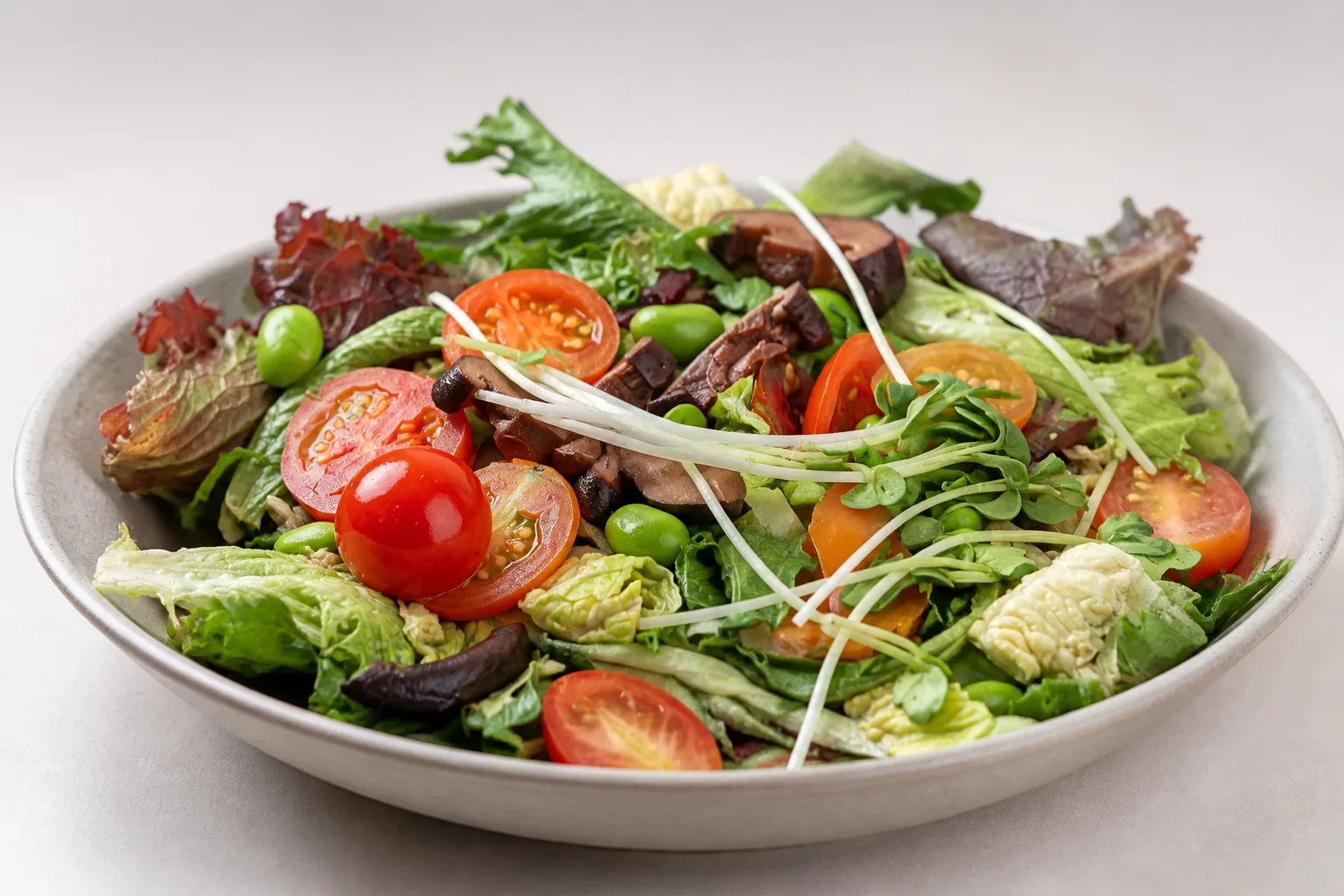 A mounded green salad with cherry tomatoes, mushrooms, edamame and sprouts in a shallow bowl, shot from a low three-quarter angle in even daylight against an empty pale background