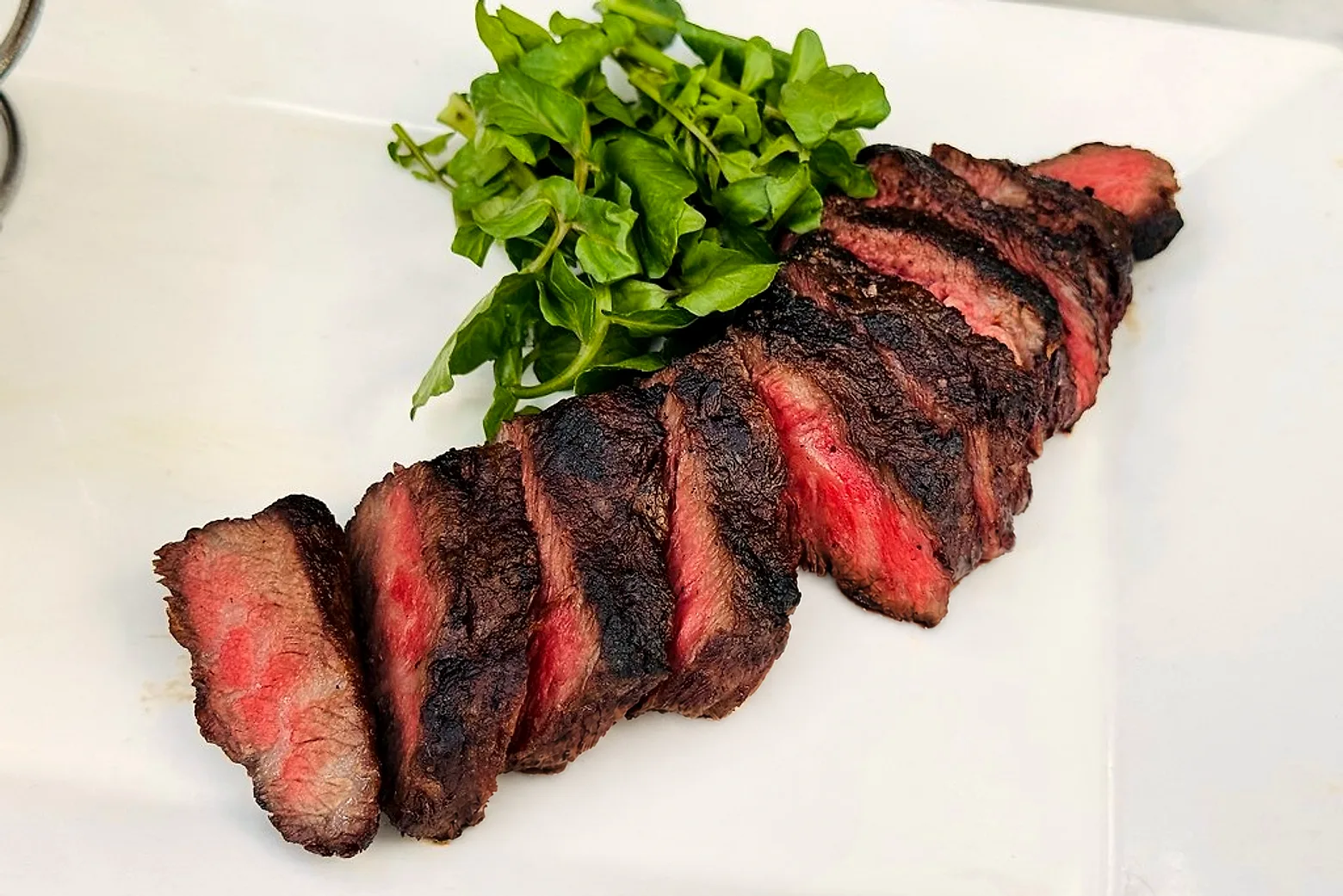 A whole grilled sirloin carved into eight thick slices and fanned diagonally across a white restaurant plate, every cut face deep red inside a heavy blackened crust, watercress tucked in behind the fan